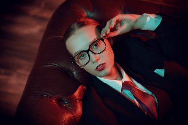 Kendine güvenen muhteşem bir kadının portresi şık bir takım elbiseyle deri bir koltukta kravat ve gözlüklerle poz veriyor. Karışık ışıklandırma. Lüks bir iş tarzı. Gözlük modası.