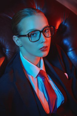 Kendine güvenen güzel bir bayan şık bir takım elbiseyle deri koltukta kravat ve gözlüklerle poz veriyor. Lüks bir iş tarzı. Gözlük modası. Karışık ışıklandırma. 