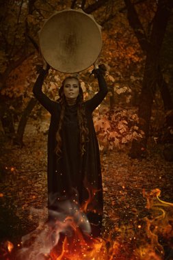 A female shaman in black garment with ethnic ornaments on her face performs a ritual with a shamanic drum in an autumn forest in front of a burning fire. Ethnic culture. Shamanic rituals. clipart