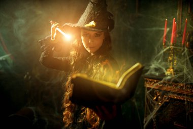 Sihirli Cadılar Bayramı büyüleri. Siyah şapkalı güzel kıvırcık saçlı büyücü sihirli bir kitap ve örümcek ağıyla kaplı eski bir kalede asasıyla büyü yapıyor. Büyücülük sanatı. 