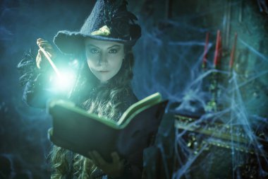 Büyücülük sanatı. Siyah şapkalı güzel kıvırcık saçlı büyücü sihirli bir kitap ve örümcek ağıyla kaplı eski bir kalede asasıyla büyü yapıyor. Sihirli Cadılar Bayramı büyüleri.