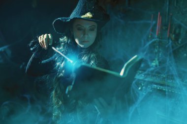 Büyücülük sanatı. Siyah şapkalı güzel kıvırcık saçlı büyücü sihirli bir kitap ve örümcek ağıyla kaplı eski bir kalede asasıyla büyü yapıyor. Sihirli Cadılar Bayramı büyüleri.