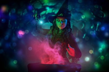 Cadılar Bayramı gecesi. Siyah elbiseli ve şapkalı güzel bir cadı karanlık bir gecede sihirli bir değnekle bir kazanı canlandırıyor. Sihir.