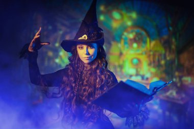 Siyah şapkalı ve siyah elbiseli güzel bir cadı sihirli bir büyü kitabıyla ortaya çıkıyor. Büyünün harika dünyası. Cadılar Bayramı. Karanlık büyülü arkaplan.
