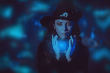 Siyah şapkalı ve elbisesi parlak bir kristal küre tutan genç cadı. Karanlık büyülü arka plan. Cadılar Bayramı büyüsü. 