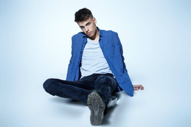 Erkek modası, gündelik tarz. Yakışıklı, esmer bir adam yerde günlük giysiler içinde poz veriyor. Beyaz bir arka planda stüdyo çekimi. Gençler. 