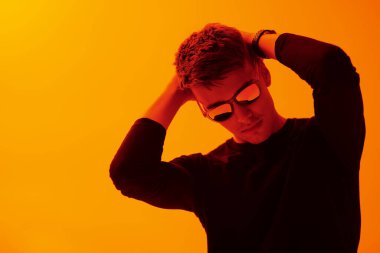 Siyah süveter ve gözlüklü yakışıklı bir gencin portresi. Kırmızı ışıkta turuncu bir stüdyoda poz veriyor. Erkek modası ve tarzı. Boşluğu kopyala.