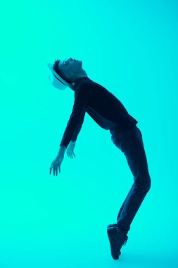 Siyah günlük giysili, güneş gözlüklü ve şapkalı parlak yeşil arka planda ayak parmaklarının üzerinde duran genç bir adamın yan görüntüsü. Moda fotoğrafı. Dans eden genç adam.