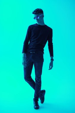 Stüdyoda poz veren siyah günlük giysili ve modaya uygun güneş gözlüklü yakışıklı bir gencin tam boy portresi. Parlak yeşil arka plan. Erkek modası, gündelik tarz..