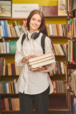 Kütüphanede elinde kitaplarla durup gülümseyen beyaz tişörtlü, modern bir liseli kızın portresi. Modern nesil. Eğitici kavram.