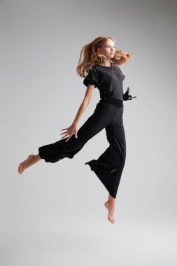 Modern bale dansları, ifadeler. Siyah bluzlu ve pantolonlu modern bir bayan dansçı gri bir stüdyo arka planına atlıyor. Tam boy portre.