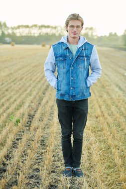 Güz tarlasında şık kotlar ve gözlük takan yakışıklı genç adam gülümsüyor. Erkek kot giyimi. 