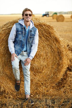 Moda giysiler ve güneş gözlüğü takan mutlu genç adam bir sonbahar tarlasında samanlığın yanında duruyor ve geniş bir şekilde gülümsüyor. Erkek kot giyimi. Gözlük stili.