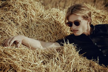 Sıcak sonbahar güneşinde samanlıkta yatan siyah gömlekli ve şık güneş gözlüklü çekici bir gencin portresi. Göz giyimi modası. Erkek güzeli..