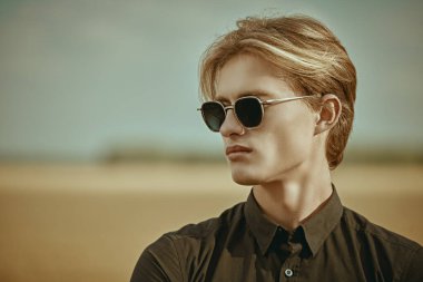 Siyah tişörtlü, sarı saçlı ve sonbahar vadisinin arka planında şık güneş gözlüklü yakışıklı bir erkek manken. Göz giyimi modası.