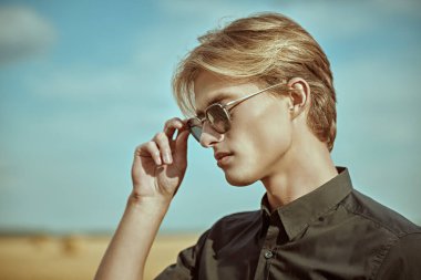 Sarı saçlı, siyah gömlekli ve şık güneş gözlüklü yakışıklı bir erkek mankenin sonbahar tarlasında ve mavi gökyüzünde yan görüntüsü. Göz giyimi modası.