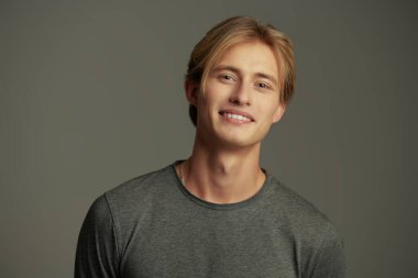 Gri stüdyo arka planında sarı saçlı, gülümseyen genç bir adamın portresi. Mutlu insanlar. Erkeklerin güzelliği ve sağlığı.