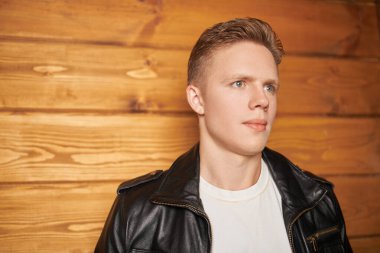 Şık deri ceketli genç sarışın adam ahşap bir duvarın yanında duruyor. Erkek modası, deri tarzı..