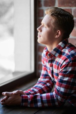 Sıradan giyinmiş genç, sarışın bir adam pencereden dikkatlice dışarı bakıyor. İnsanlar, yaşam tarzı.