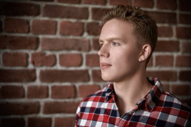 Kareli gömlekli yakışıklı modern bir adam tavan arası bir odada tuğla bir duvarın yanında duruyor. Gençler, yaşam tarzı.. 