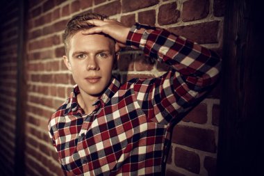 Kareli gömlekli yakışıklı modern bir adam tavan arası bir odada tuğla bir duvarın yanında duruyor. Gençler, yaşam tarzı. Sıradan erkek modası..