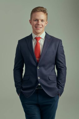 İş adamı. Takım elbiseli, beyaz gömlekli ve kırmızı kravatlı başarılı bir gencin portresi. Cebinde el ele tutuşup neşeyle gülümsüyor. Gri arka plan. Başarılı insanlar..