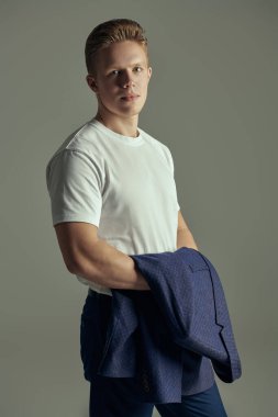 Beyaz tişört ve pantolon giyen yakışıklı, modern bir genç adam elinde ceketiyle ayakta duruyor. Gri arka plan. Gençler. Erkek modası, gündelik tarz..