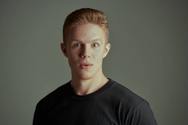 Siyah tişörtlü yakışıklı bir sarışının portresi kameraya şaşkınlık ve heyecanla bakıyor. Gri arka plan. Gençler, duygular. 