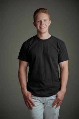 Kameraya gülümseyen siyah tişörtlü ve beyaz pantolonlu yakışıklı bir adamın stüdyo fotoğrafı. Gri arka plan. Gençler. Günlük erkek modası, gençlik tarzı..