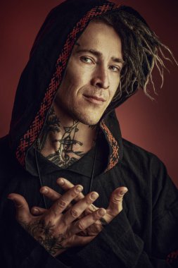 Siyah etnik kıyafetli yetişkin bir erkek şaman kafasında bir başlıkla kameraya gülümsüyor. Koyu kırmızı arka plan. Şamanizm. Cadılar Bayramı, Fantezi.