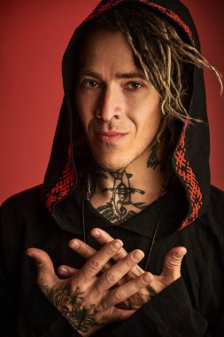 Etnik başlıklı giyinmiş dövmeleri ve rastaları olan modern bir erkek şaman kameraya sakince bakıyor. Koyu kırmızı arka plan. Şamanizm. Cadılar Bayramı, Fantezi.