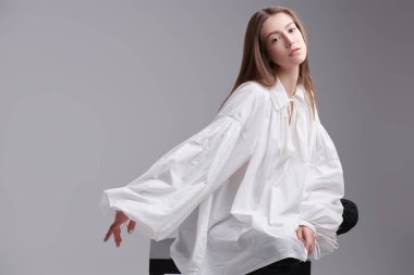 Kadın güzelliği ve sağlığı. Taze tenli, güzel kahverengi saçlı ve geniş kollu beyaz gömlekle poz veren güzel bir kız resmi. Gri arkaplan. 