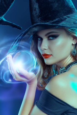 Cadılar Bayramı 'nda sihir. Siyah şapkalı ve zarif makyajlı siyah elbiseli çekici genç cadı parlayan bir kristal küre tutuyor. Koyu mavi arka plan. Parti posteri. 
