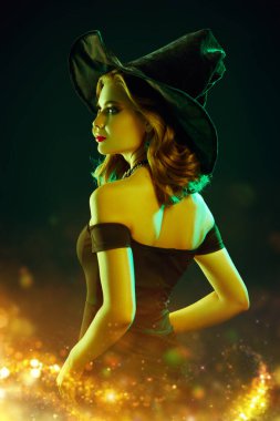 Siyah şapkalı çekici bir cadı ve şık makyajlı siyah elbisesi sihirli ışıklarla siyah bir arka planda duruyor. Parti posteri. 