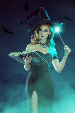 Siyah şapkalı güzel genç bir cadı büyü kitabı ve sihirli değnekle büyü yapıyor. Gizemli dumanla çevrili. Uçan yarasaların olduğu koyu mavi bir arka plan. Cadılar Bayramı partisi posteri. 