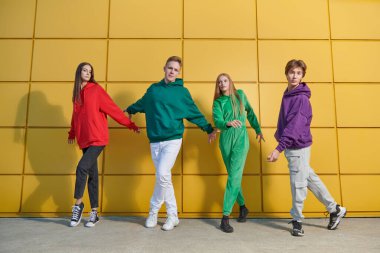 Modern gençlik kıyafetleri içinde dört aktif genç açık sarı bir binanın yanında sokak balesi yapıyor ve birlikte eğleniyorlar. Parlak gençlik modası. Modern gençler işte. Sokak dansları.