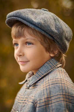 Sonbahar parkında kareli ceketli ve şapkalı gülümseyen sarışın bir çocuğun portresi. Çocuk modası. Çocuklar ve duygular.