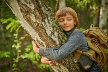 Eko-turizm. Sırt çantalı mutlu bir çocuk yaz ormanında duruyor ve neşeyle bir ağaca sarılıyor. Macera ve seyahat tutkusu.