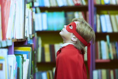 Okul üniforması giymiş ve süper kahraman kırmızı pelerinli kütüphanede kitap seçen bir çocuğun yan görüntüsü. Eğitimde süper yetenekler. Çocukların rüyaları. 