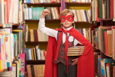 Okul üniformalı, süper kahraman kırmızı pelerinli ve maskeli bir çocuk kütüphanede elinde kitaplarla yumruğunu kaldırıyor. Eğitimde süper yetenekler. Çocukların rüyaları. 
