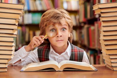 Düzgün üniformalı zeki bir öğrenci, bir okul kütüphanesinde kitap yığınlarının arasında büyüteçle ilginç bir şekilde ansiklopedi çalışıyor. Modern eğitim ve çocuklar.