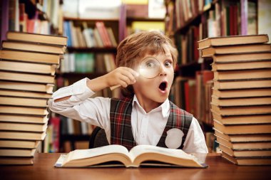Düzgün üniformalı zeki bir öğrenci, bir okul kütüphanesinde kitap yığınlarının arasında büyüteçten kameraya merakla bakıyor. Modern eğitim ve çocuklar.