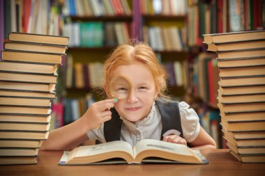 Komik bir liseli kız kütüphanede kitap yığınlarının arasında oturmuş büyüteçle kameraya gülümsüyor. Modern çocuklar ve eğitim.