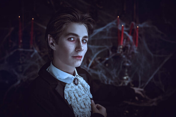 Halloween. An attractive young vampire in an elegant tailcoat stands by the fireplace in an old castle covered in cobwebs and looks at the camera calmly. Vampires novel. Copy space.