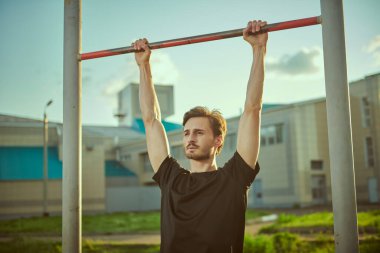 Sportif genç bir adam parktaki açık hava eğitimi sırasında yatay bir barda asılı duruyor. Spor açık havada. Aktif ve sağlıklı yaşam tarzı.