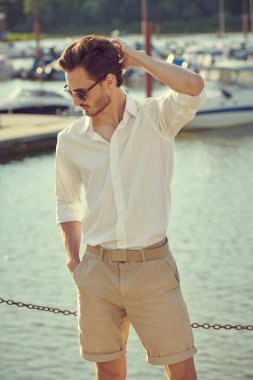 Yat limanında beyaz gömlekli, şortlu ve siyah güneş gözlüklü çekici bir adamın portresi. Erkek güzelliği ve moda. Denizde yaz tatili.