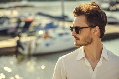 Yat limanında zarif bir gömlek ve siyah güneş gözlüğü takmış koyu renk saçlı bir adam duruyor. Deniz yolculukları. Yaz tatili. Erkek yaşam tarzı. Zarif erkek modası ve tarzı. Boşluğu kopyala.