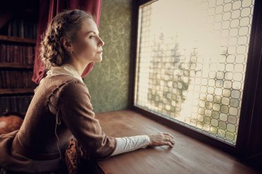 18. yüzyıl İskoç elbisesi giymiş güzel bir aristokrat kız klasik bir iç mimariye sahip bir odada düşünceli bir şekilde pencereden dışarı bakar. Romanın kadın kahramanı. 18. yüzyıl tarzı.