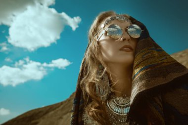 Boho tarzı, yaz tarzı. Kafasında etnik başörtüsü olan zarif boho tarzı mücevherler içinde güzel bir kadın kum tepelerinin ve mavi gökyüzünün arka planında duruyor. Boşluğu kopyala.