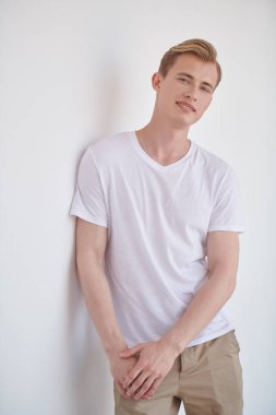 Erkek güzelliği ve sağlığı. Yaz kıyafetleri içinde yakışıklı genç bir sarışın beyaz bir duvara yaslanmış duruyor. Gençler. 